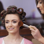 Makeup artist applying blush with brush during women's salon makeup session with hair styling preparation